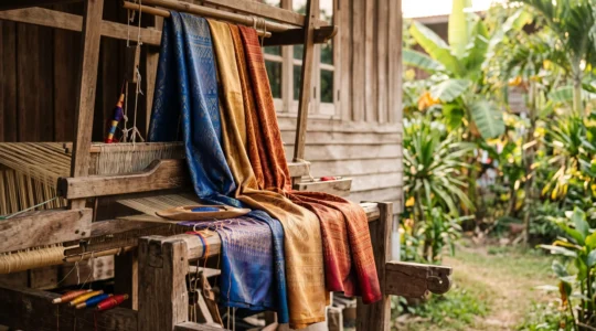 Écharpes de soie thaïlandaise aux couleurs chatoyantes disposées sur un métier à tisser traditionnel en bois, éclairées par une lumière naturelle chaude